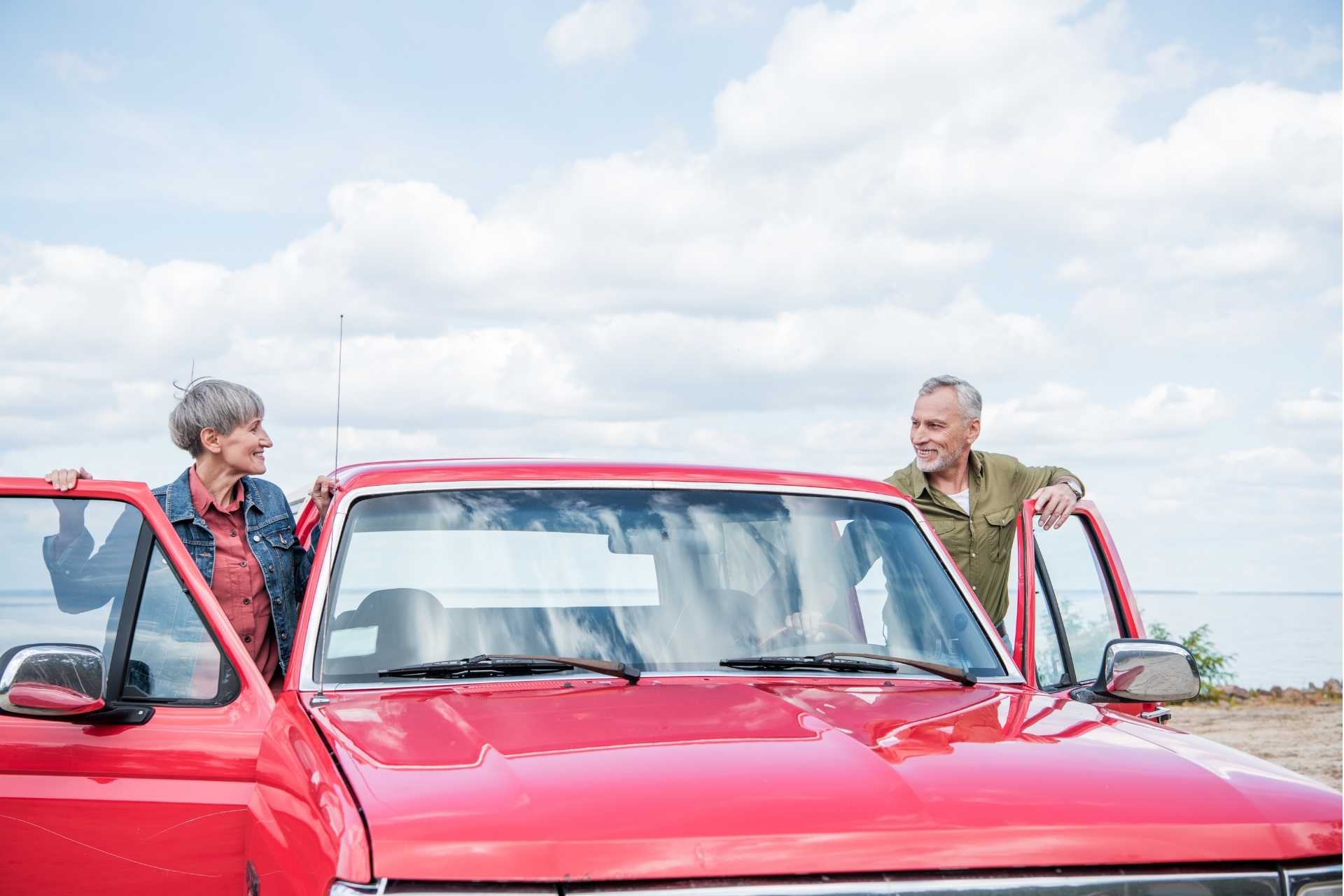 Senior couple smiling at each other while getting into a red classic car.