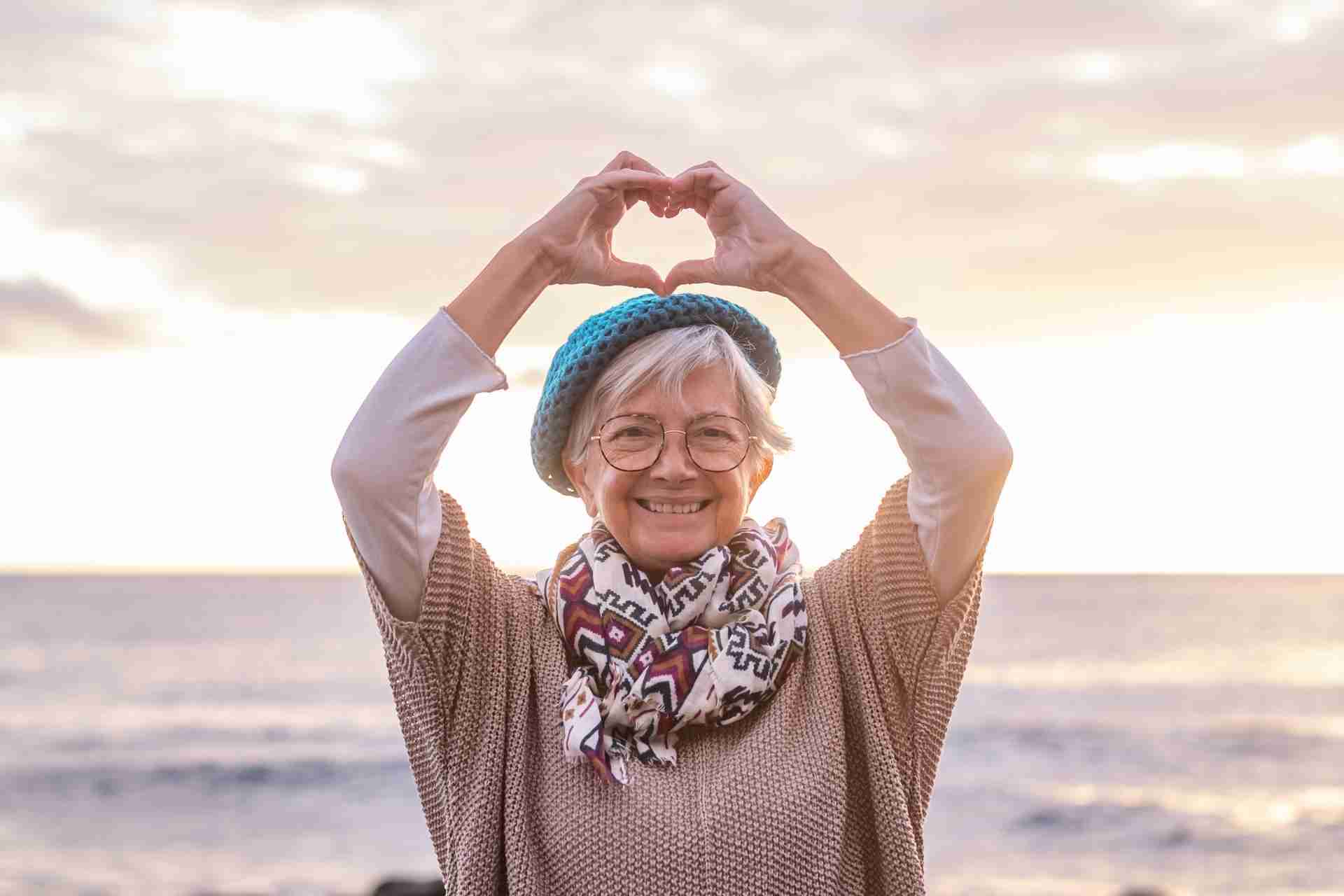 A senior woman on the beach holding her hands over her head in a heart shape.